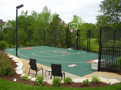 Outdoor multi-sport court with green flooring and white markings with s basketball hoop surrounded by black fencing.