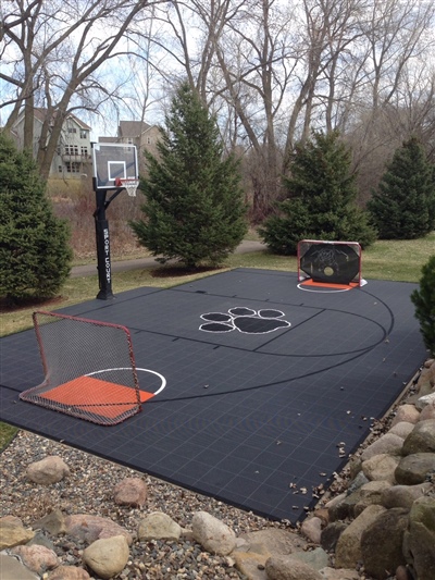 An outdoor black sport court with a basketball hoop, hockey goals, and surrounded by trees and a rock border.