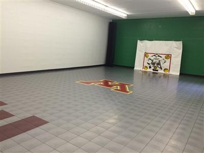 Indoor sports court with grey flooring, a hockey goal, and a central U of M logo. 