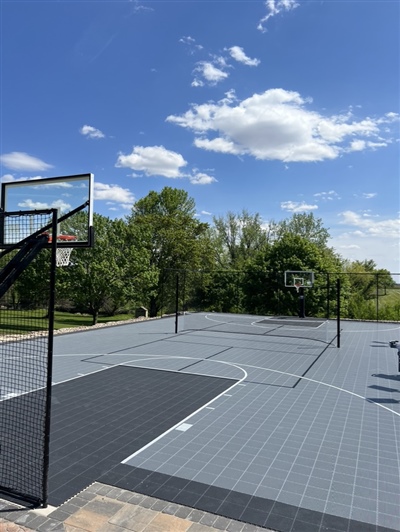 Outdoor residential sports court with a light and dark grey surface, featuring a basketball hoop and surrounded by trees and greenery.
