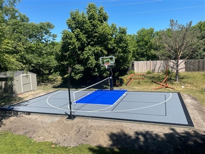 Outdoor residential sports court with a gray surface and a blue painted key area, featuring a basketball hoop and tennis net, and surrounded by trees and a fence in Fox Point.