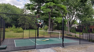 Outdoor residential sports court with a black and green surface, featuring a basketball hoop and a tennis net, surrounded by a black fence, trees, and greenery.