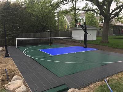 Outdoor residential sports court with a green and black surface, a blue painted key area, a basketball hoop, a volleyball net, and surround by a wooden fence in Elm Grove.
