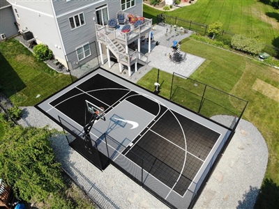 Outdoor residential sports court with a black and gray surface, featuring a basketball hoop and central court logo, and surrounded by a black fence.