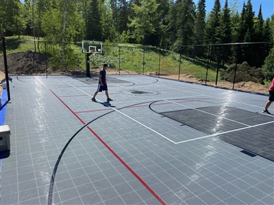 Outdoor residential sports court with a gray surface and red lines, surrounded by trees and greenery.
