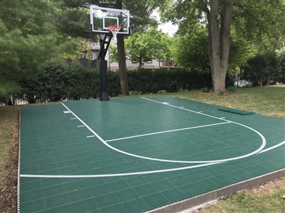 A basketball court with a green surface, white lines, and a basketball hoop in a wooded backyard.