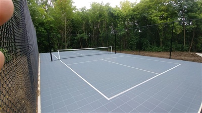 Outdoor sports court with a gray surface, white lines, and a tennis net, surrounded by trees and greenery.