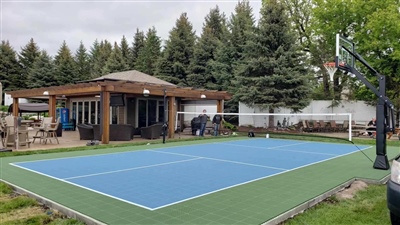 Outdoor residential sports court with a blue and green surface, a basketball hoop, and a nearby wooden pergola, surrounded by trees and greenery.